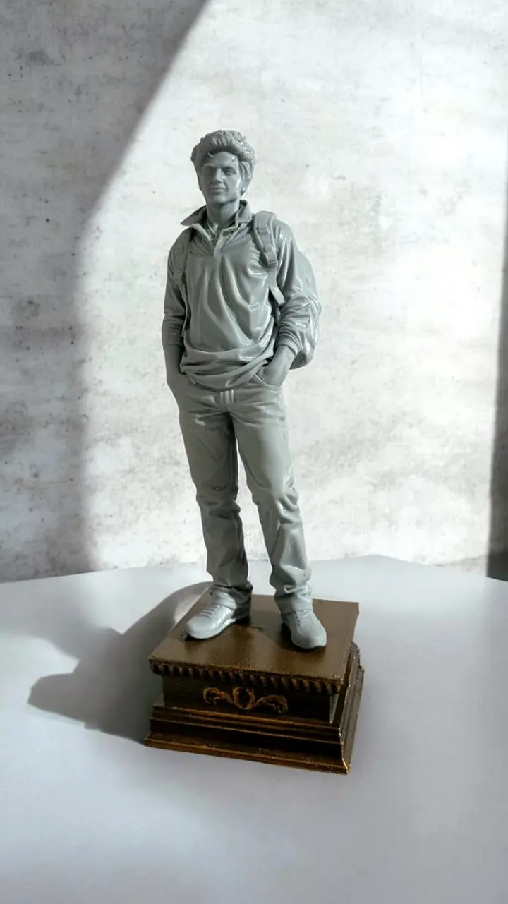 Estátua impressa em 3D de São Carlo Acutis, jovem com mochila e tênis, em acabamento cinza sobre base de madeira