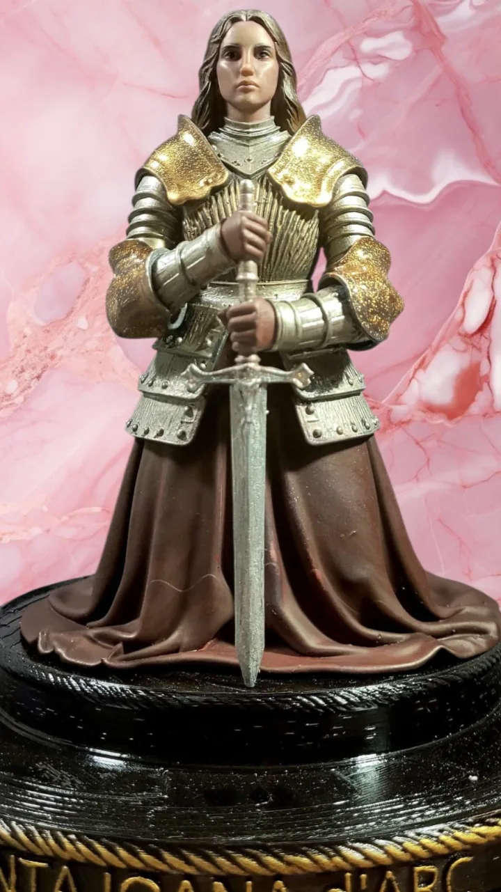 Imagem policromada de Santa Joana d'Arc ajoelhada em armadura prateada com detalhes dourados, com as mãos apoiadas na espada, sobre base escura com o nome da santa