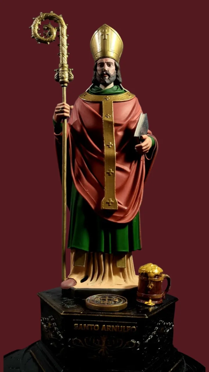 Imagem policromada de Santo Arnulfo com mitra e báculo dourados, casula vermelha e verde, com caneca de cerveja e medalha junto à base, sobre pedestal escuro com o nome do santo