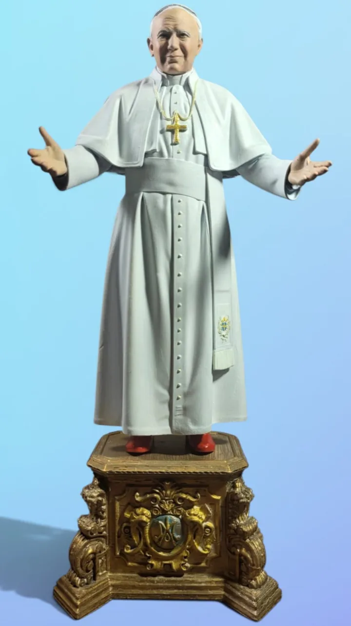 Estátua em 3D policromada de São João Paulo II de braços abertos, em vestes brancas com cruz peitoral dourada e sapatos vermelhos, sobre base ornamentada dourada