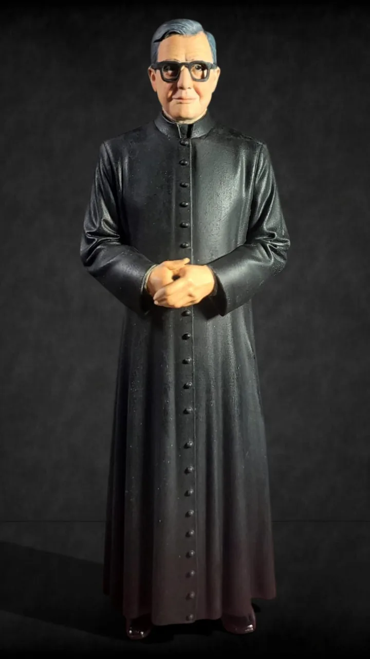 Estátua em 3D policromada de São Josemaria Escrivá em batina preta, de óculos e mãos unidas, sobre base escura